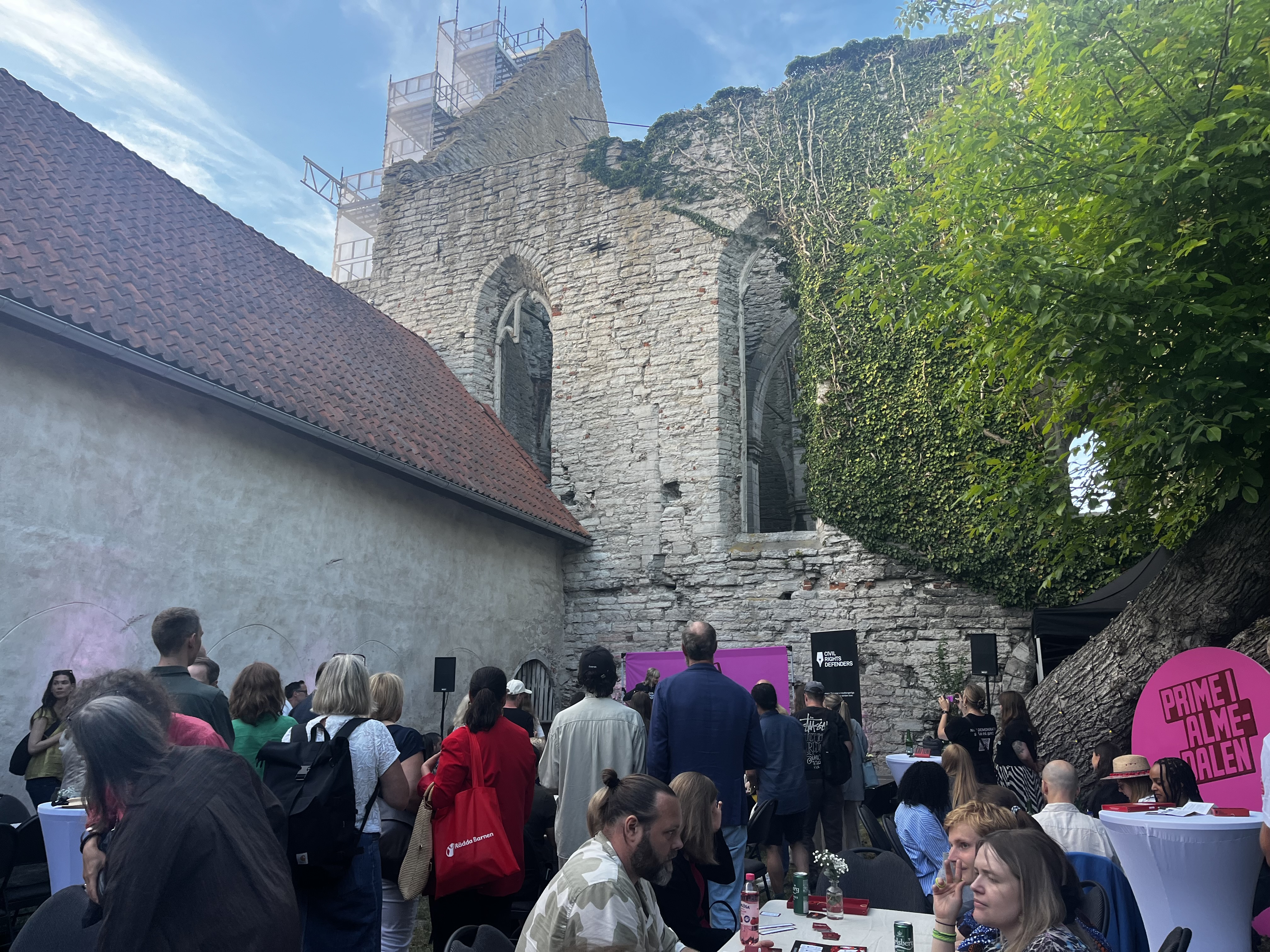 Människor samlas vid en utomhusfest framför en gammal ruinstuga, med en scen och dekorationer i bakgrunden.