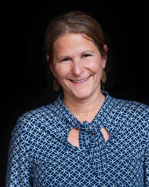 Kvinna i en mönstrad blus med knytning, framför en mörk bakgrund.