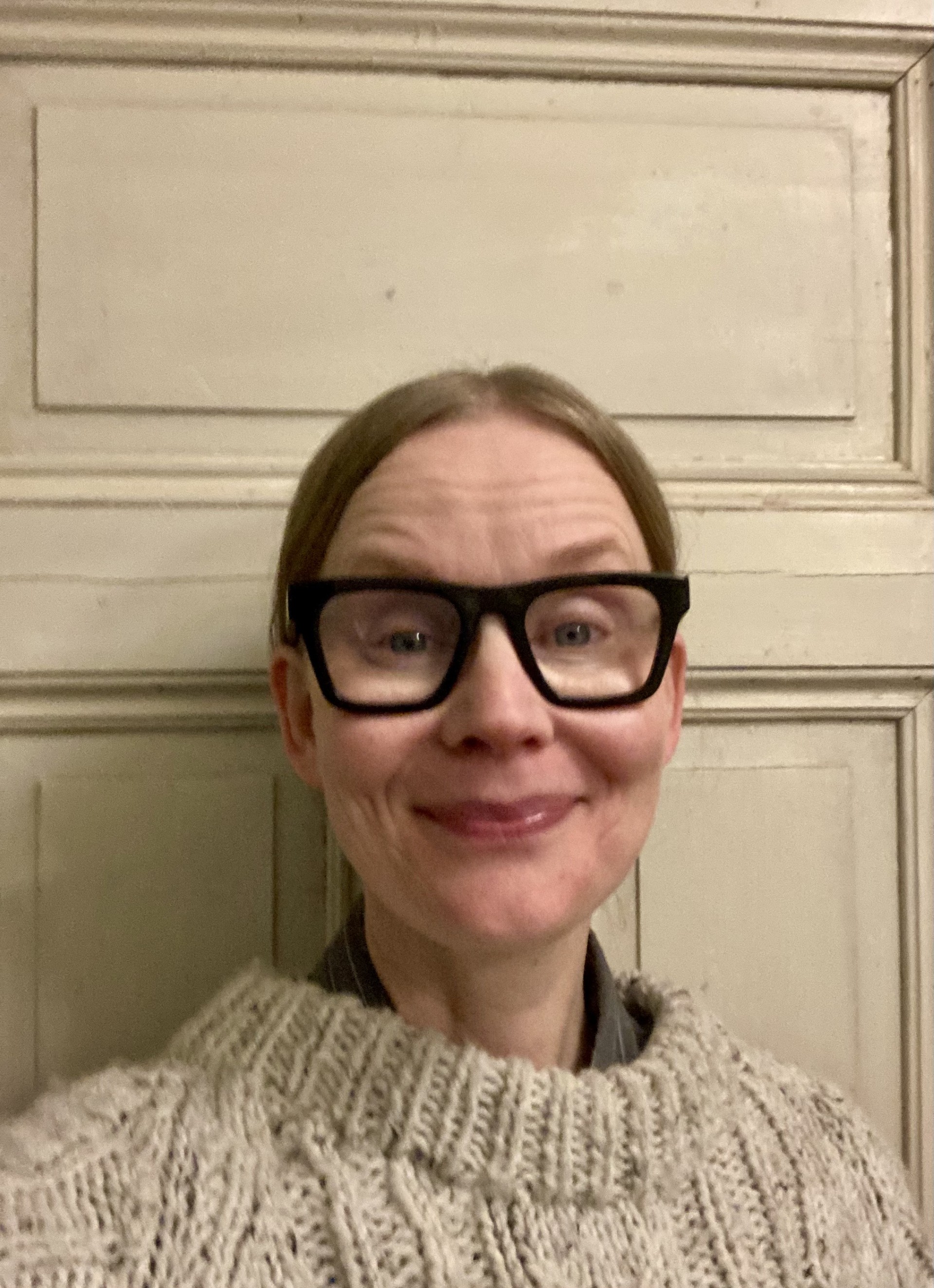 Person i stickad tröja står framför en ljus vägg med dörrpaneler, troligtvis i ett hem.