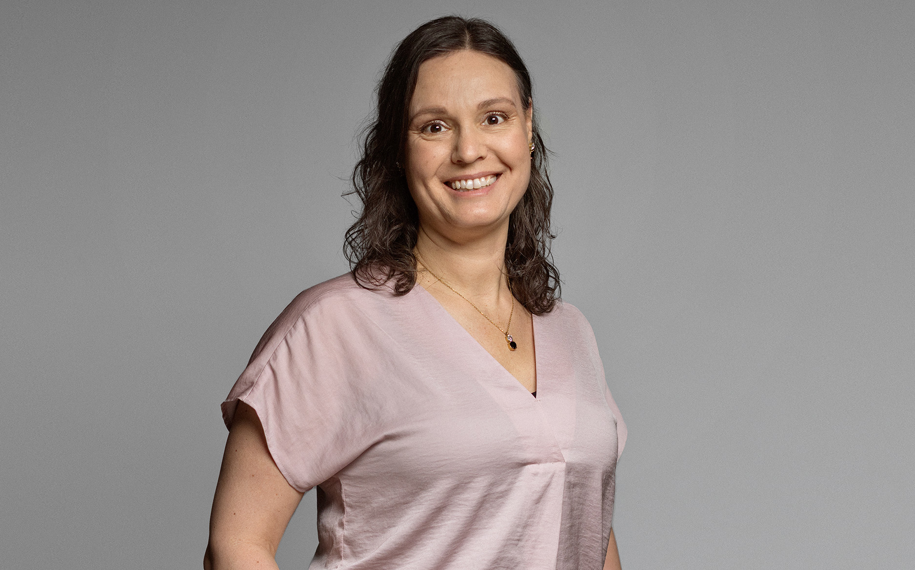 Kvinna i ljusrosa blus står framför en grå bakgrund, med en avslappnad hållning.